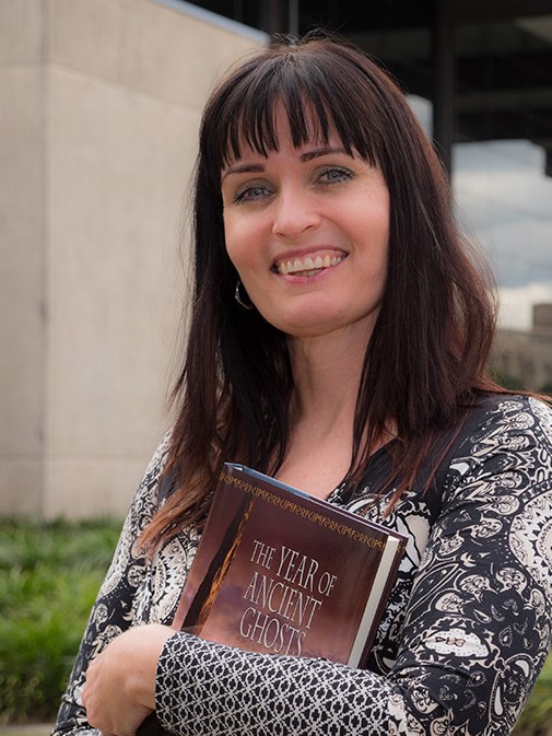 Professor Kim Wilkins, a white woman with long dark brown hair, smiles at the camera. She is holding a book titled "The Year of Ancient Ghosts."