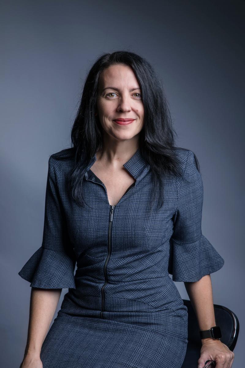 Associate Professor Nicole Hartley, a white woman with long black hair, is smiling at the camera. She is wearing a dark grey business dress.