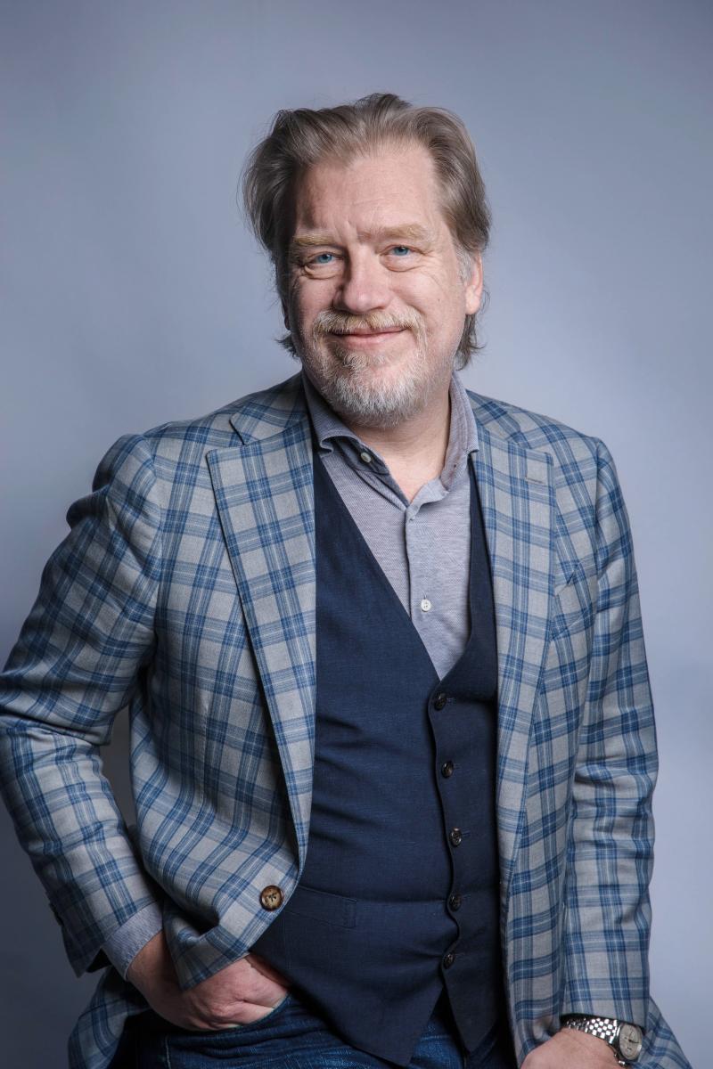 Associate Professor Tim Kastelle, a white man with ginger hair and grey beard, is smiling at the camera. He is wearing a grey shirt, navy waistcoat, and grey and navy checked jacket.