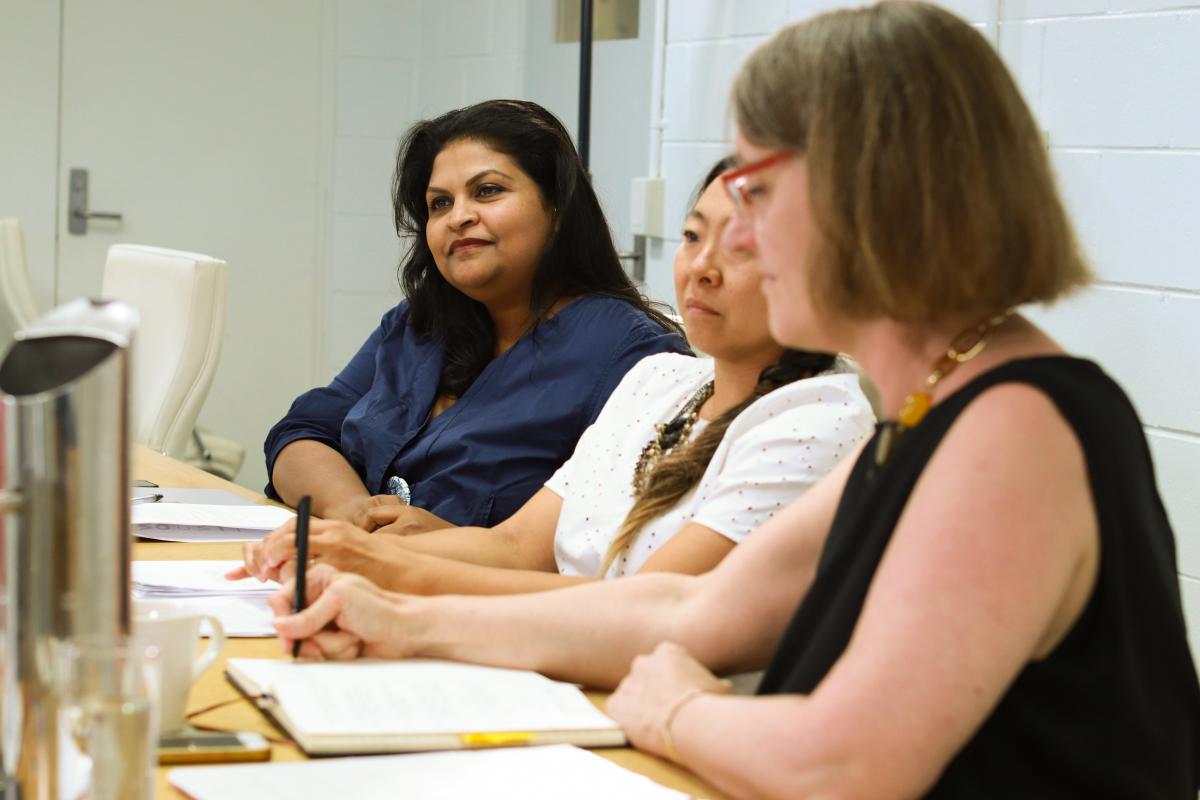 Three culturally diverse women listening to someone (out of frame). Dr Kate Power is taking notes.