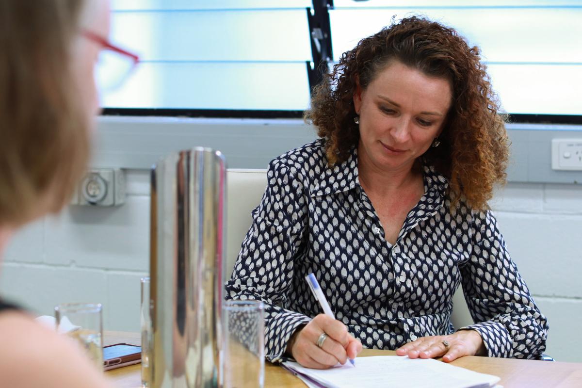 White woman with curly brown hair, wearing a black and white spotted shirt, is taking notes. 